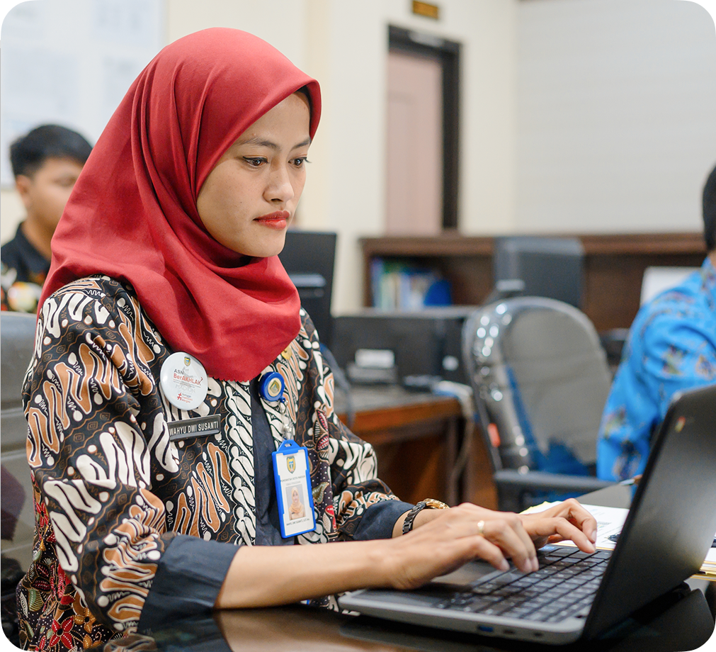 Seorang perempuan mengenakan hijab sedang bekerja menggunakan perangkat Chromebook di ruang kelas.