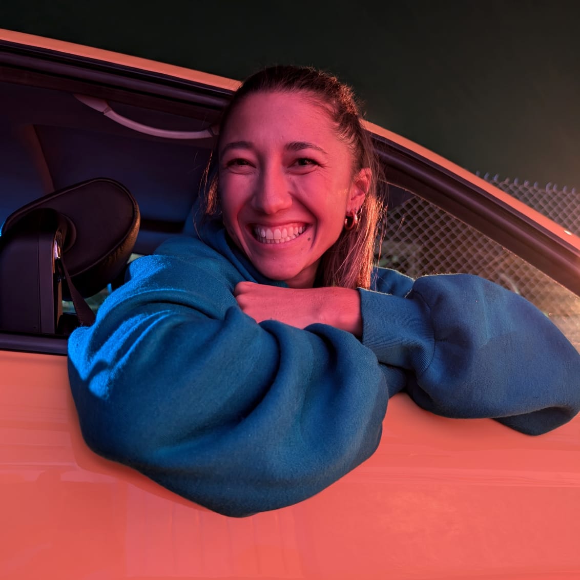 A person leans out of a car, smiling, in dim pink light.